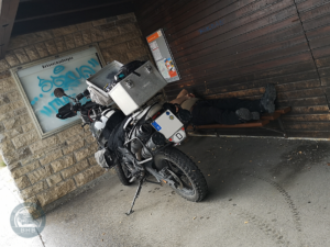 Der Road Captain liegt auf einer Bank in einem Bushaltestellenwartehäuschen. Mit unter dem Dach steht die BMW F650 GS.